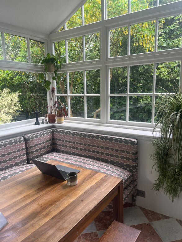 Dining area with wooden table and patterned bench in a sunroom with large windows. starring Georgie corner banquette