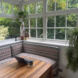 Dining area with wooden table and patterned bench in a sunroom with large windows. starring Georgie corner banquette