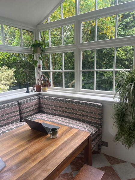 Dining area with wooden table and patterned bench in a sunroom with large windows. starring Georgie corner banquette
