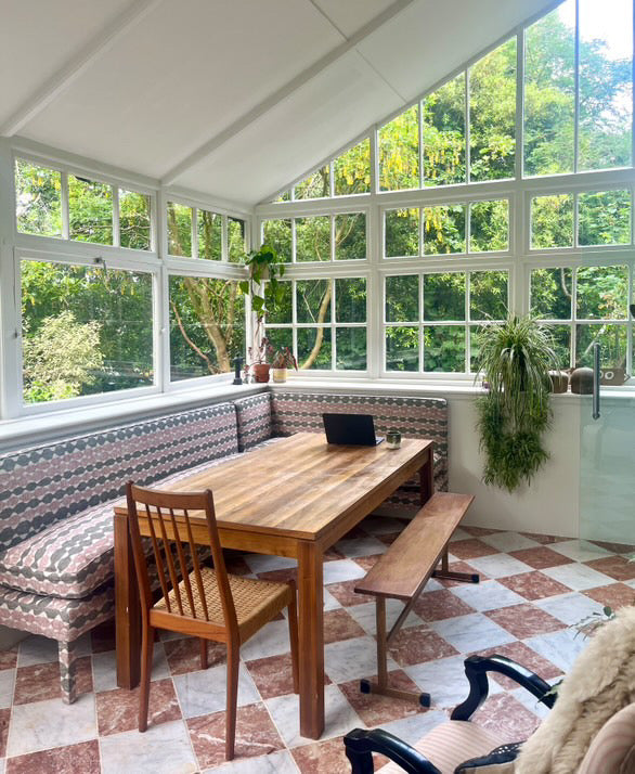 Dining area with wooden table and chairs in a room with large windows and greenery outside. starring geode corner banquette