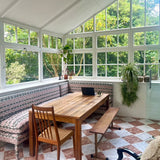 Dining area with wooden table and chairs in a room with large windows and greenery outside. starring geode corner banquette