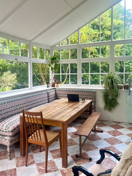 Dining area with wooden table and chairs in a room with large windows and greenery outside. starring geode corner banquette