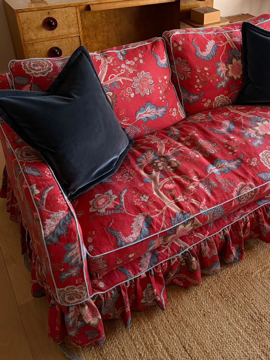 A red floral velvet slipcover with a ruffled skirt and light blue piping, styled on a sofa with two black velvet pillows in a warm indoor setting.