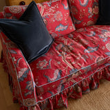 A red floral velvet slipcover with a ruffled skirt and light blue piping, styled on a sofa with two black velvet pillows in a warm indoor setting.