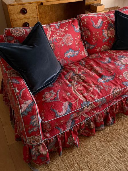 A red floral velvet slipcover with a ruffled skirt and light blue piping, styled on a sofa with two black velvet pillows in a warm indoor setting.