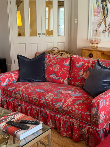 A red floral ruffled sofa slipcover with two dark navy velvet throw pillows, styled in a cozy living room setting.
