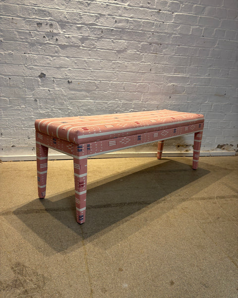 A rectangular upholstered bench with pink and white geometric patterned fabric, set against a white brick wall and concrete floor.