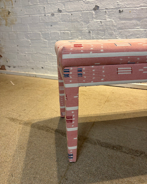 A close-up of a pink upholstered table leg and corner, showing a pink, white, and blue geometric patterned fabric, set against a white brick wall and concrete floor.