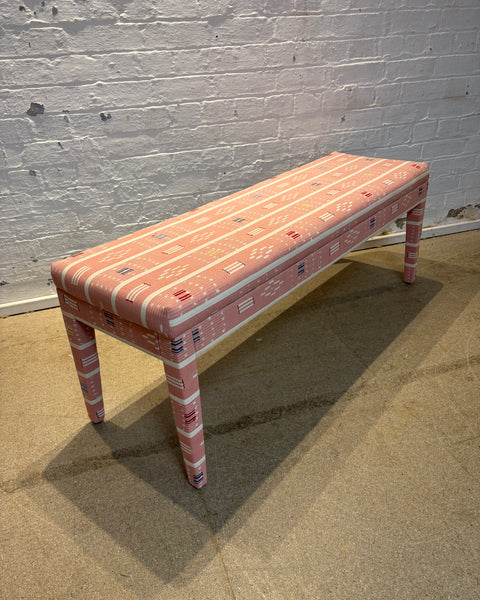 A rectangular upholstered bench with a pink and white geometric pattern, positioned in front of a white brick wall on a concrete floor.