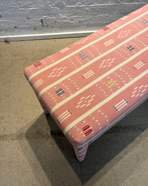 A rectangular upholstered bench with a pink and white geometric patterned fabric, shown in a warehouse setting with a concrete floor and white brick wall.