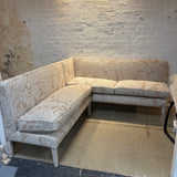 An L-shaped corner sofa upholstered in a beige and white damask patterned fabric, set against a white brick wall with a cardboard floor protector.