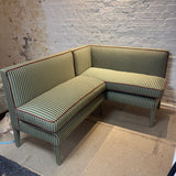 An L-shaped corner sectional sofa upholstered in green and beige vertically striped fabric with thin red piping along the edges, set against a white brick wall.