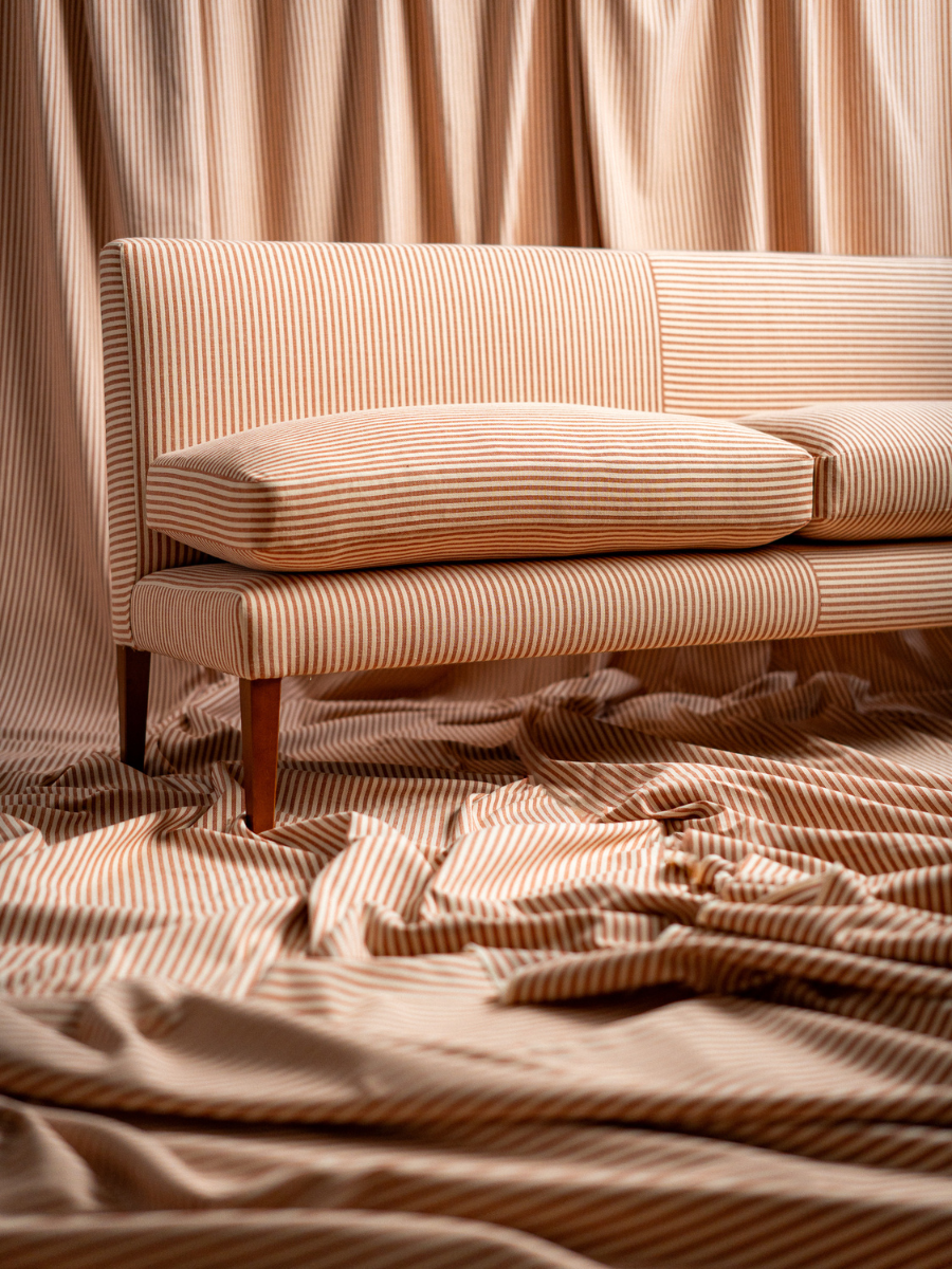 A beige upholstered sofa with thin vertical brown stripes, two matching seat cushions, and dark wooden legs, styled in a bedroom-like setting with matching striped bedding and draped fabric in the background.